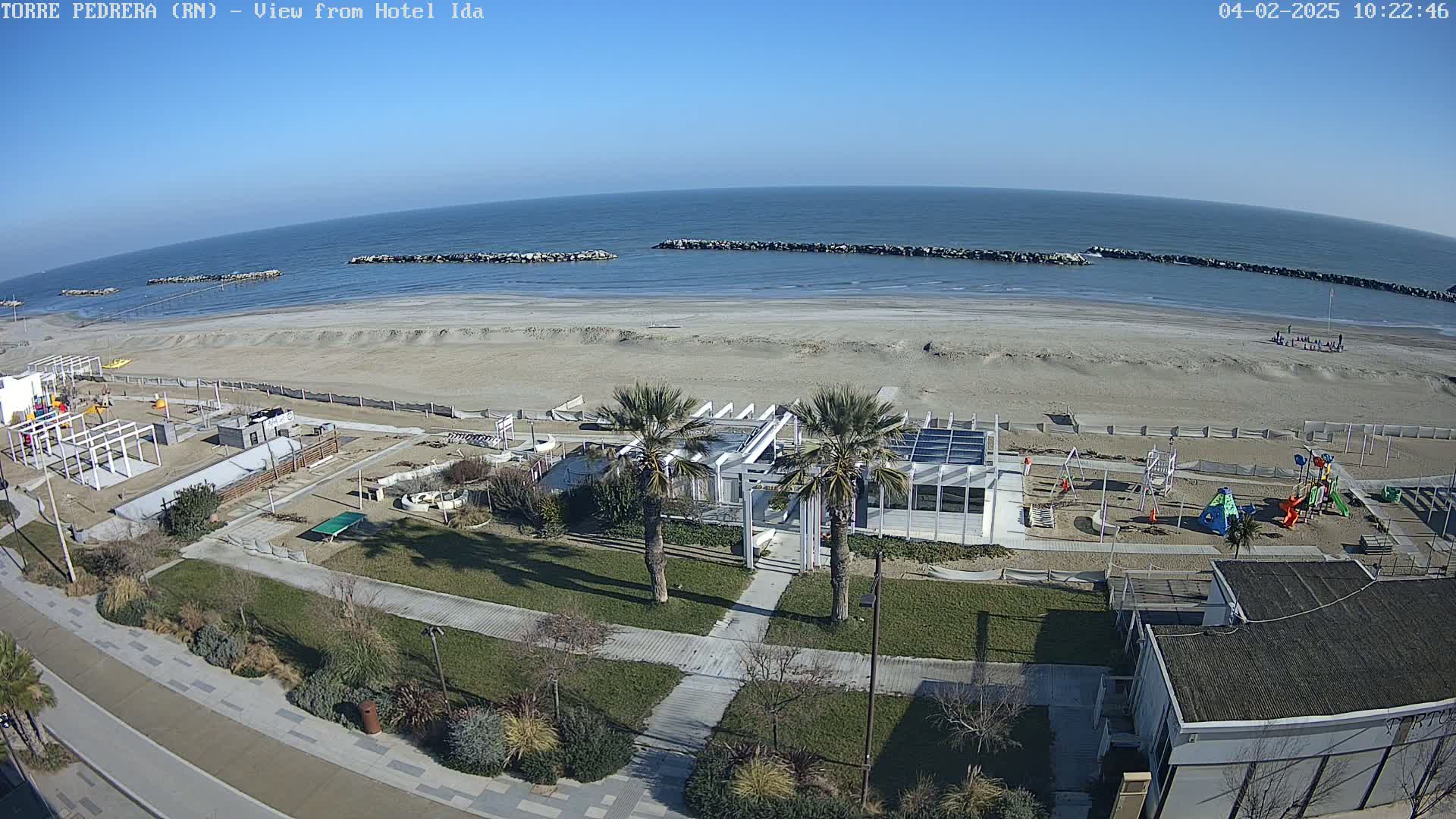 A sandy beach with calm ocean waters under a clear blue sky features a playground, a beachfront building with palm trees, and a paved walkway leading to the shore.