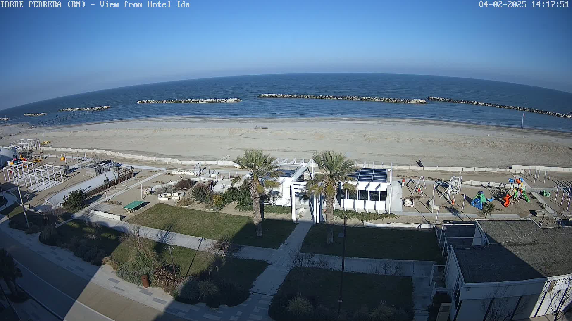 A sunny beach with calm ocean water, a playground, and a modern building is seen from an elevated vantage point.