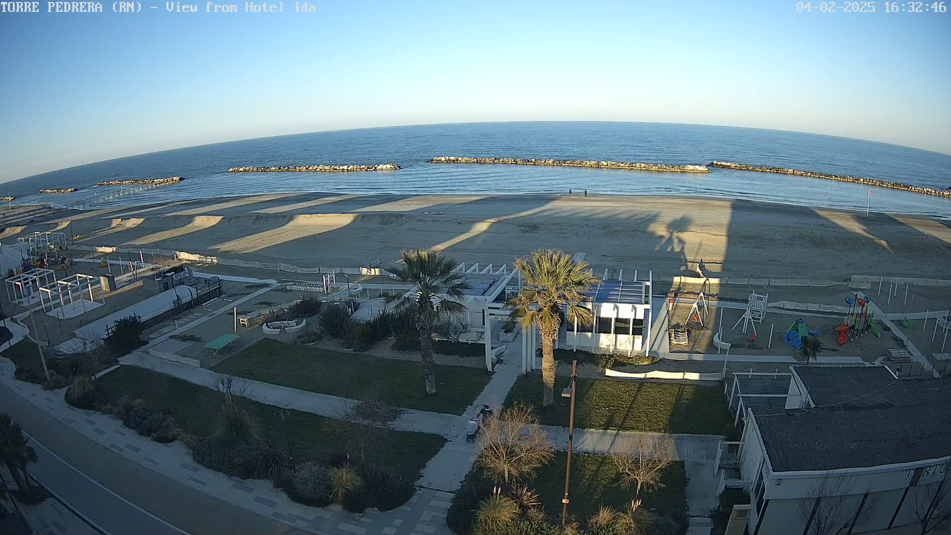 A beach with calm, blue water under a clear, bright sky features a playground, beachfront buildings, and palm trees casting long shadows on the sand.