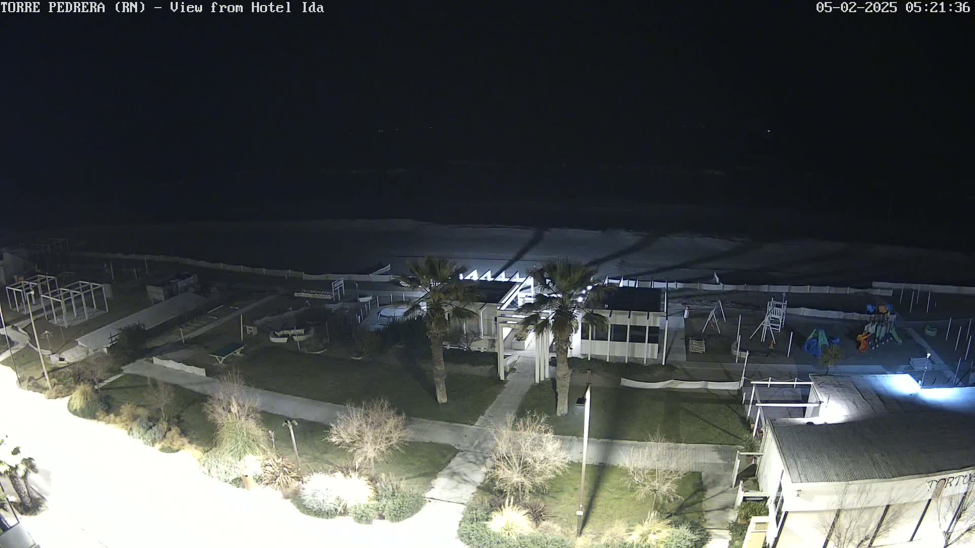 A nighttime aerial view shows a beach, a building with palm trees, a playground, and other structures under a dark sky.