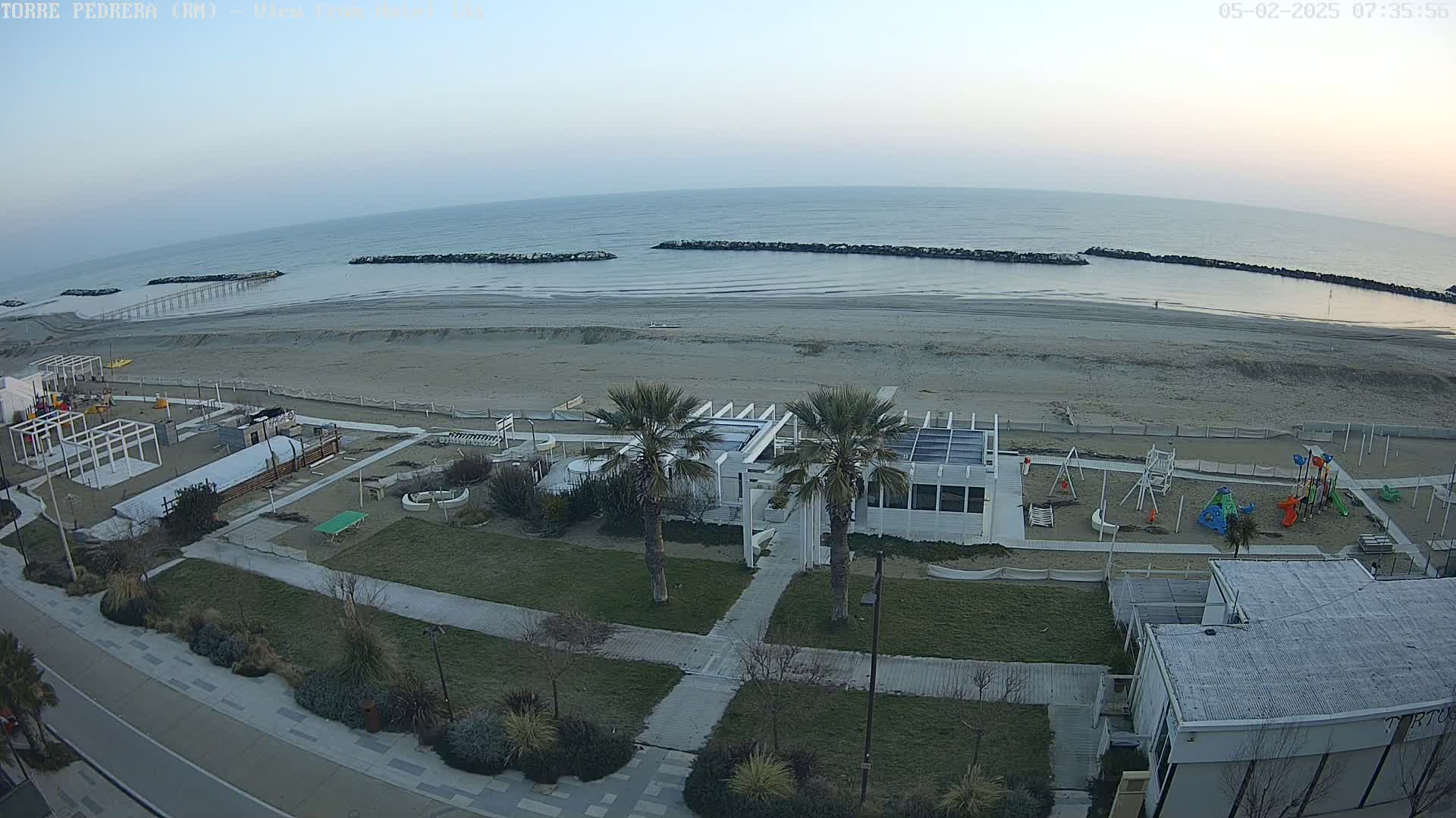 A beach with a calm sea under a hazy sky, featuring a beachfront building with palm trees, a playground, and a walking path.