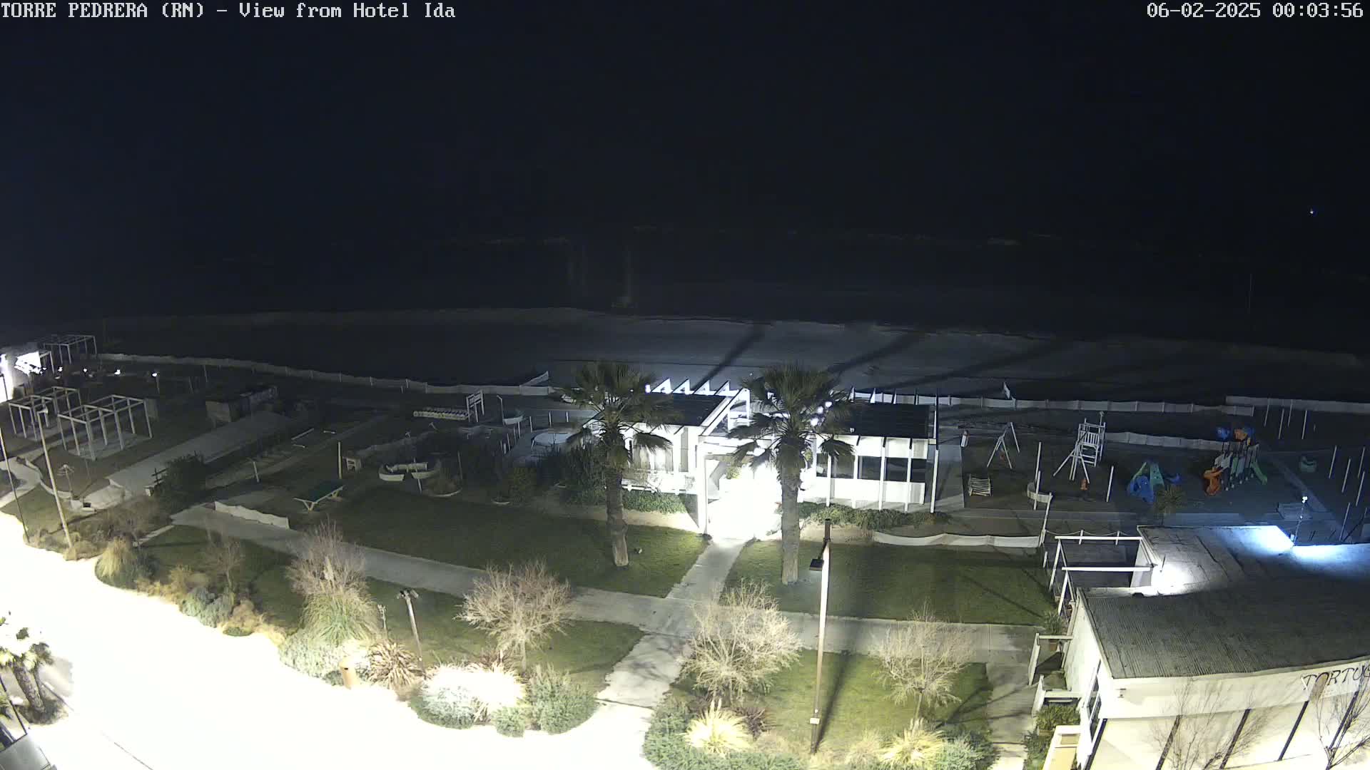 A nighttime aerial view shows a beach, a playground, a building with palm trees in front, and other structures, all illuminated under dark skies.