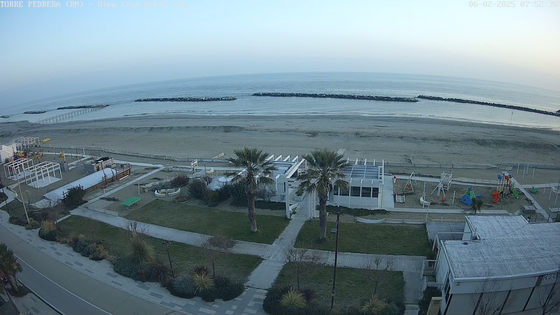 A beach with a calm sea under a clear, light-grey sky, features a playground, beachfront buildings, and palm trees.