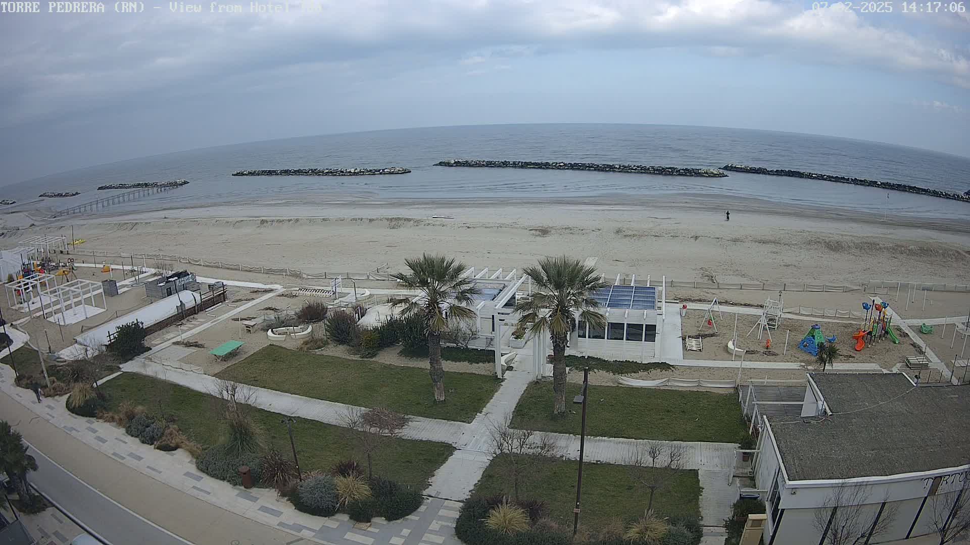 An overcast day at a beach shows a sandy shore, calm sea with breakwaters, a playground, a white beachside building, and palm trees, all viewed from above.