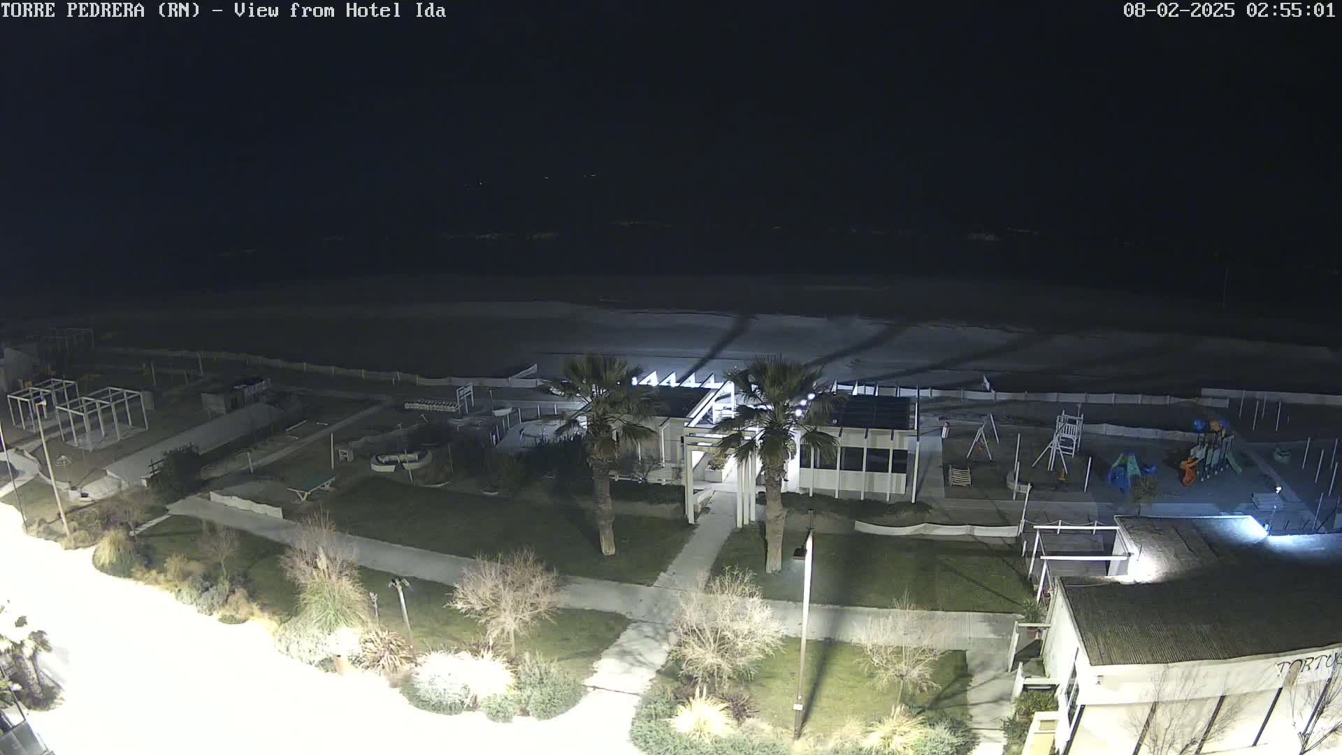 A nighttime aerial view shows a beach, a playground, and a building with palm trees, all illuminated by artificial light.