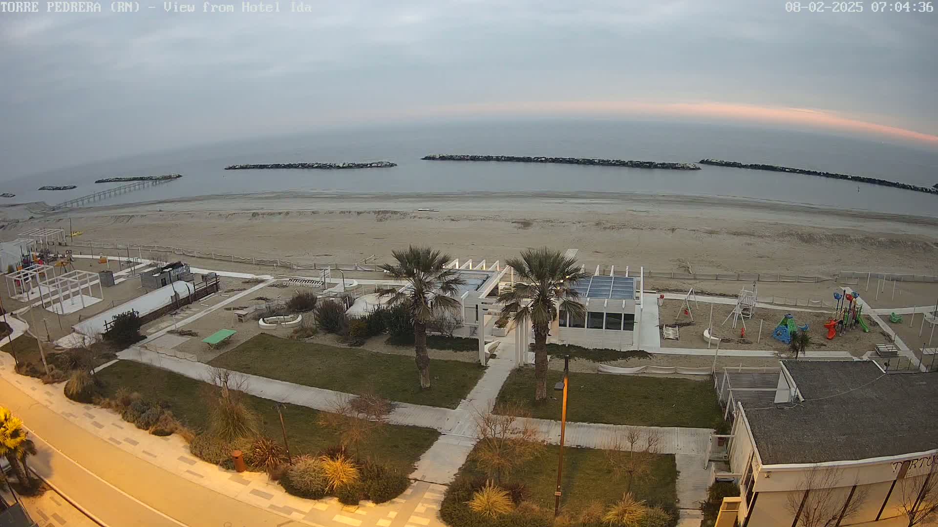 A sandy beach with a playground, palm trees, and a white building is visible, under an overcast sky with a hint of sunrise or sunset.
