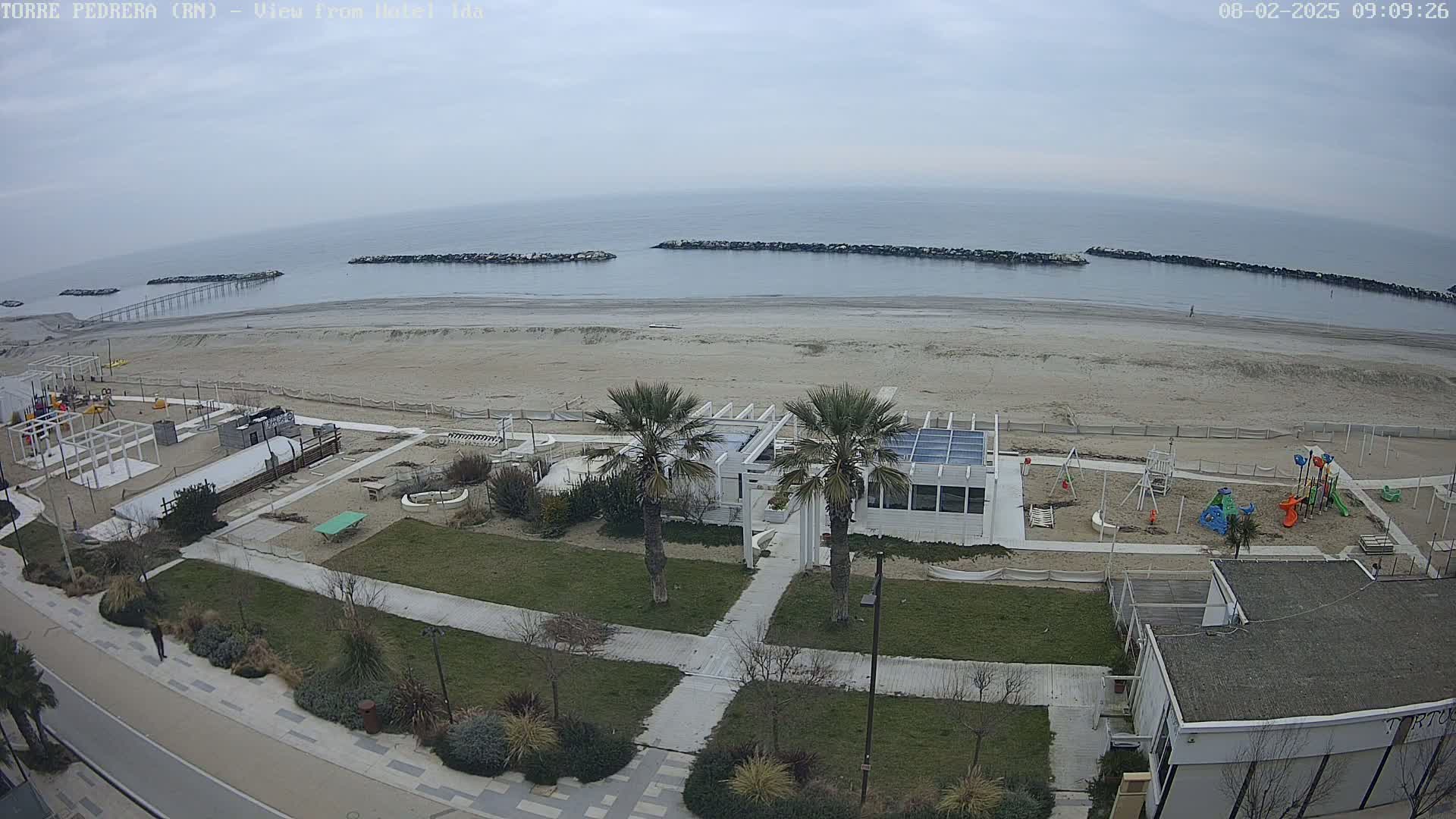An overcast day shows a beach with a playground, beachfront buildings, and palm trees, all viewed from above.