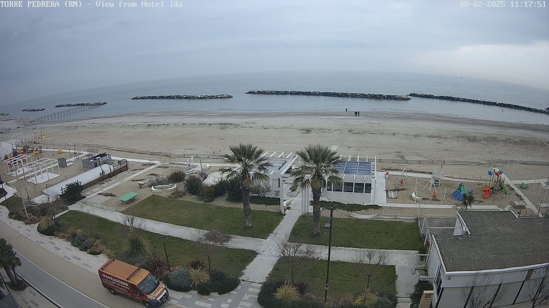 An overhead view of a beach on an overcast day shows a sandy shoreline, calm ocean water, breakwaters, a playground, a small white building, and palm trees.
