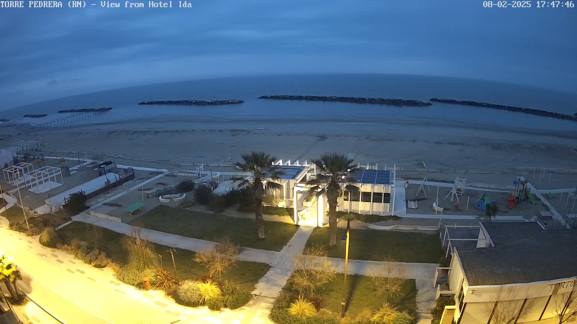 An aerial view at dusk shows a beach with a playground, a modern building, palm trees, and a road leading to it, all under a cloudy sky.