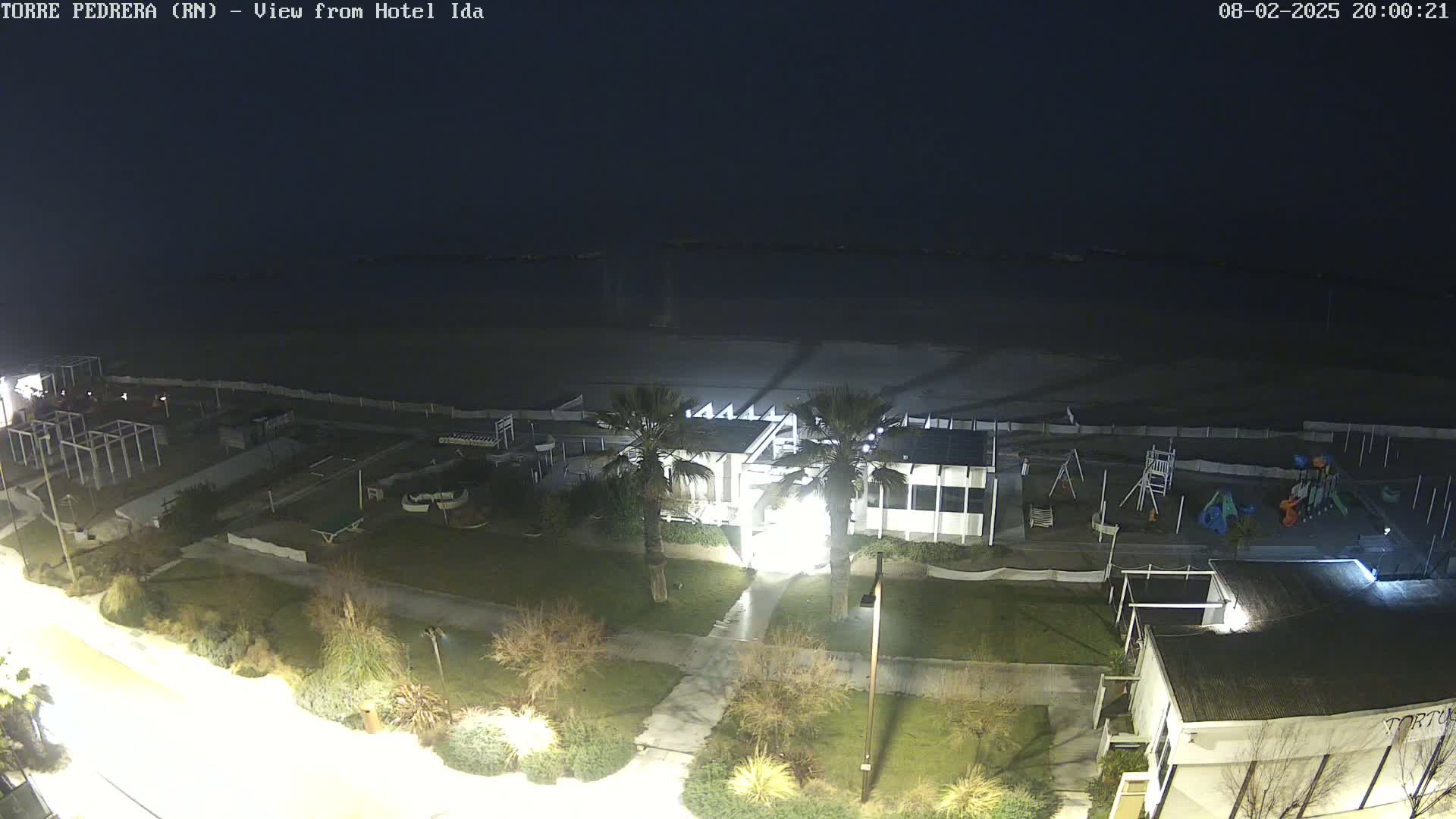 A nighttime aerial view of a beach, showing a playground, beachfront building, and palm trees under a dark sky.