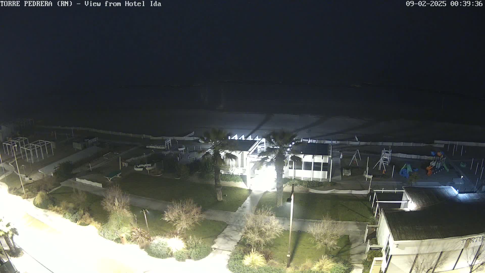 A nighttime aerial view of a beach area showing a playground, some buildings, palm trees, and a dark ocean under a clear, dark sky.