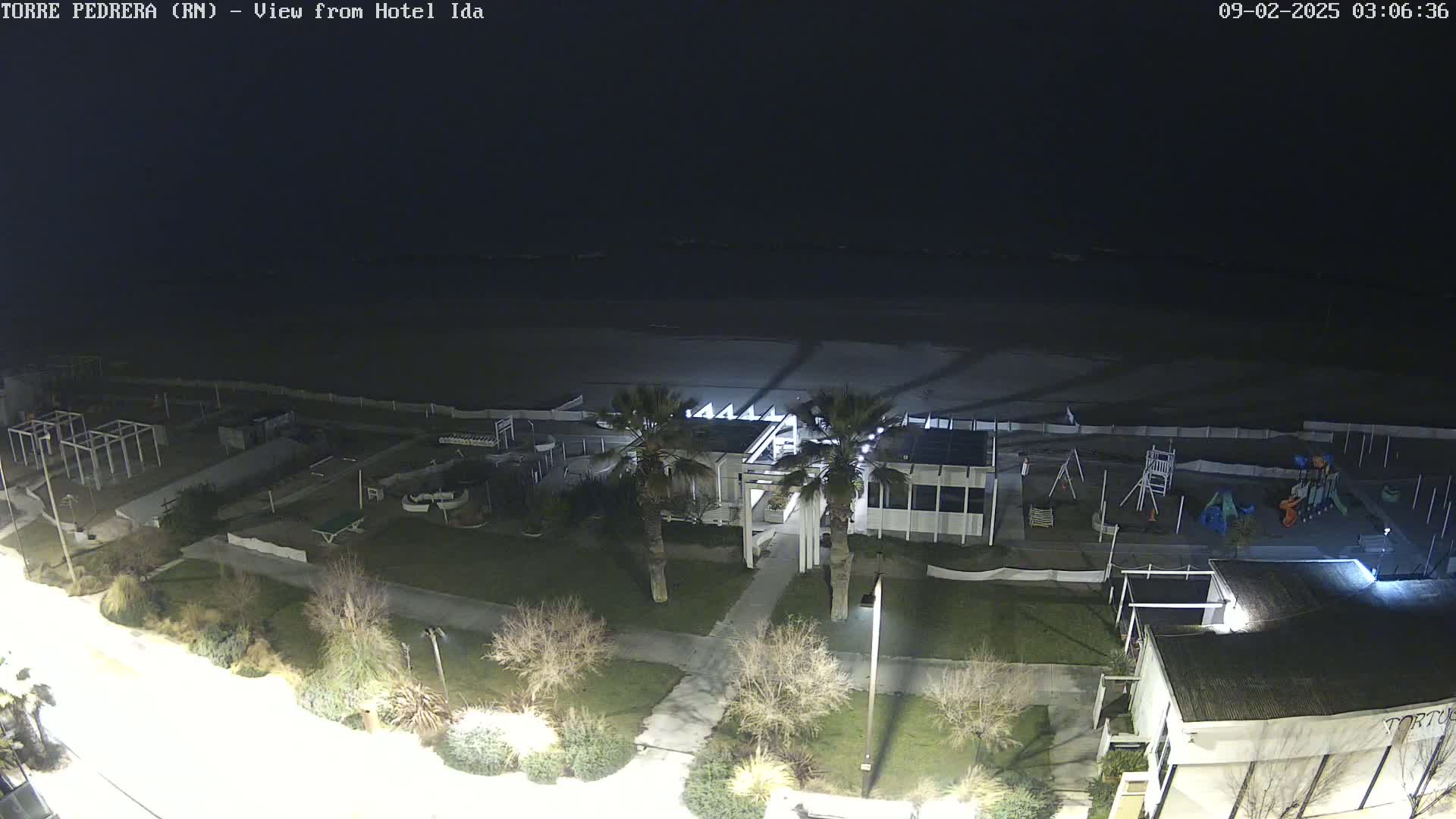 A nighttime aerial view shows a beach, playground, and buildings with palm trees under a dark sky.