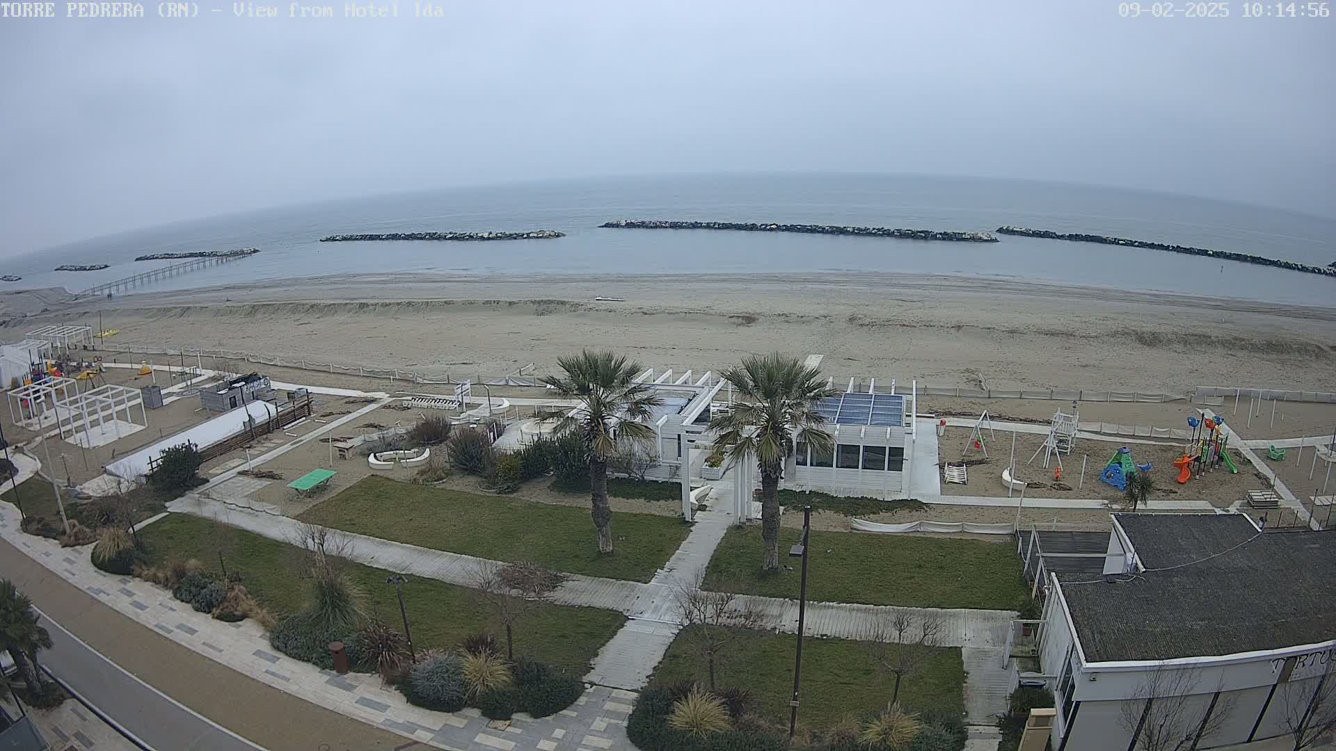 An overcast day reveals a beach scene with a sandy shore, calm water, breakwaters, a playground, a white building, palm trees, and a paved walkway.