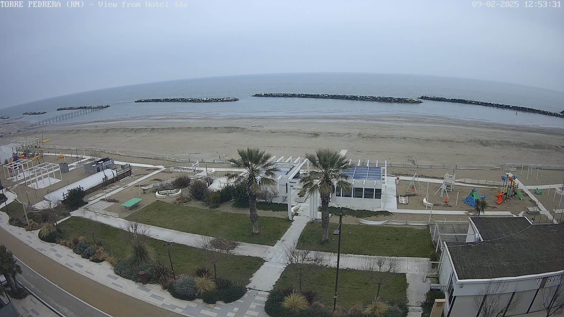 An overcast day shows a beach with a playground, a white building, palm trees, and a paved walkway next to the ocean, which is protected by rock barriers.