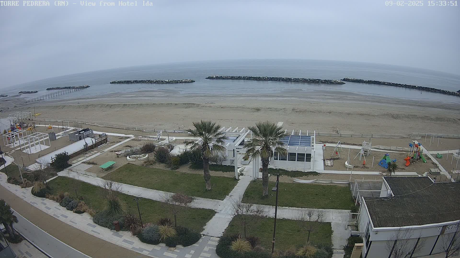 A beach scene on an overcast day shows a sandy beach, calm ocean, a white building with palm trees, a playground, and a paved walkway.