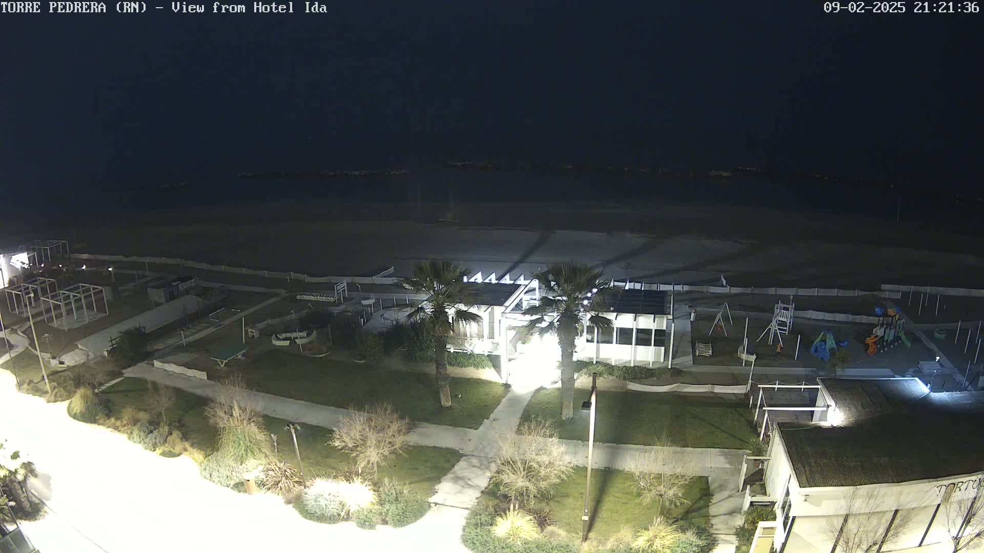 A nighttime, aerial view of a beach with a building, playground, and palm trees under a dark sky.