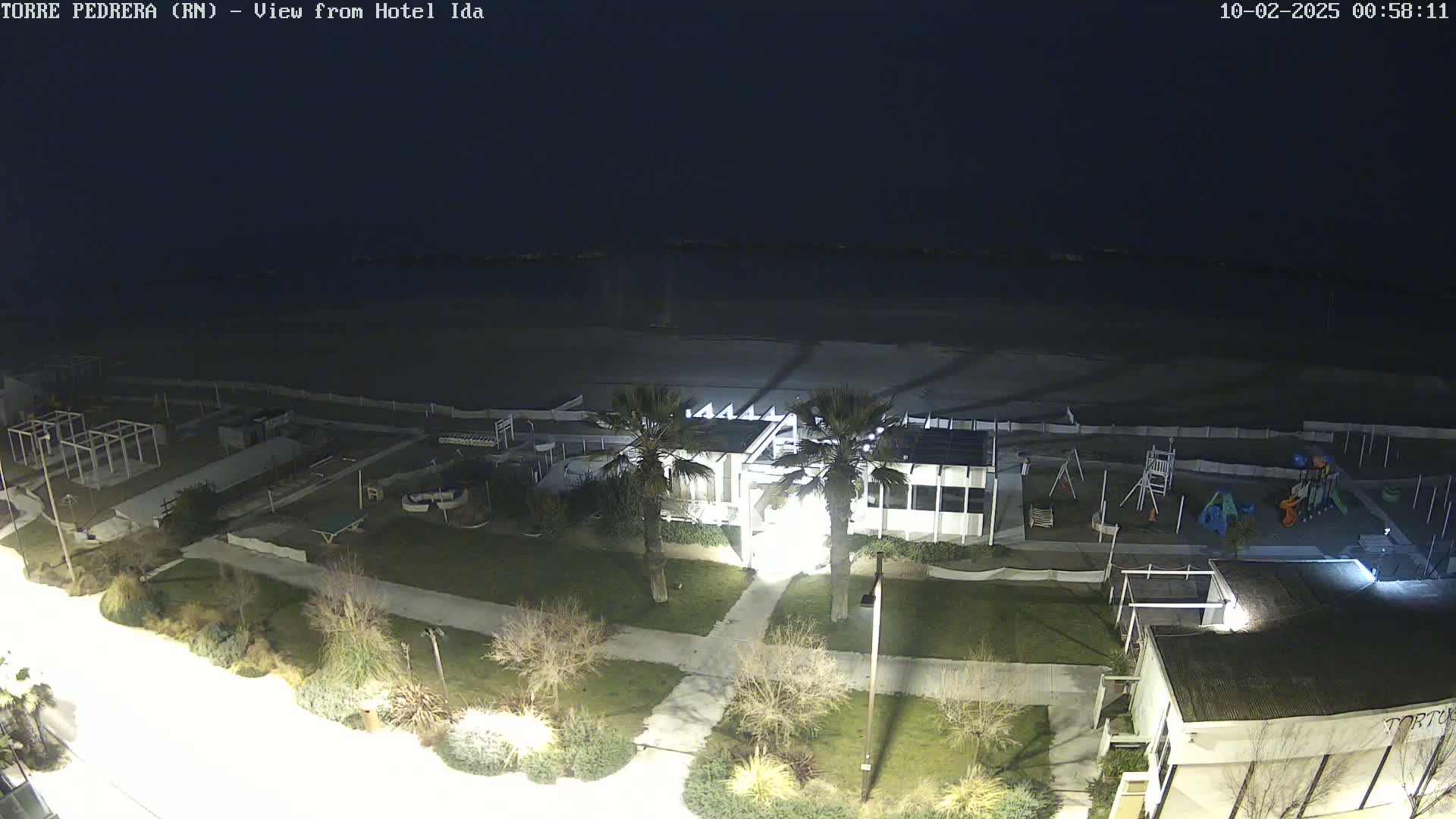 A nighttime aerial view shows a beach, a playground, a building with palm trees, and other structures illuminated by artificial light.