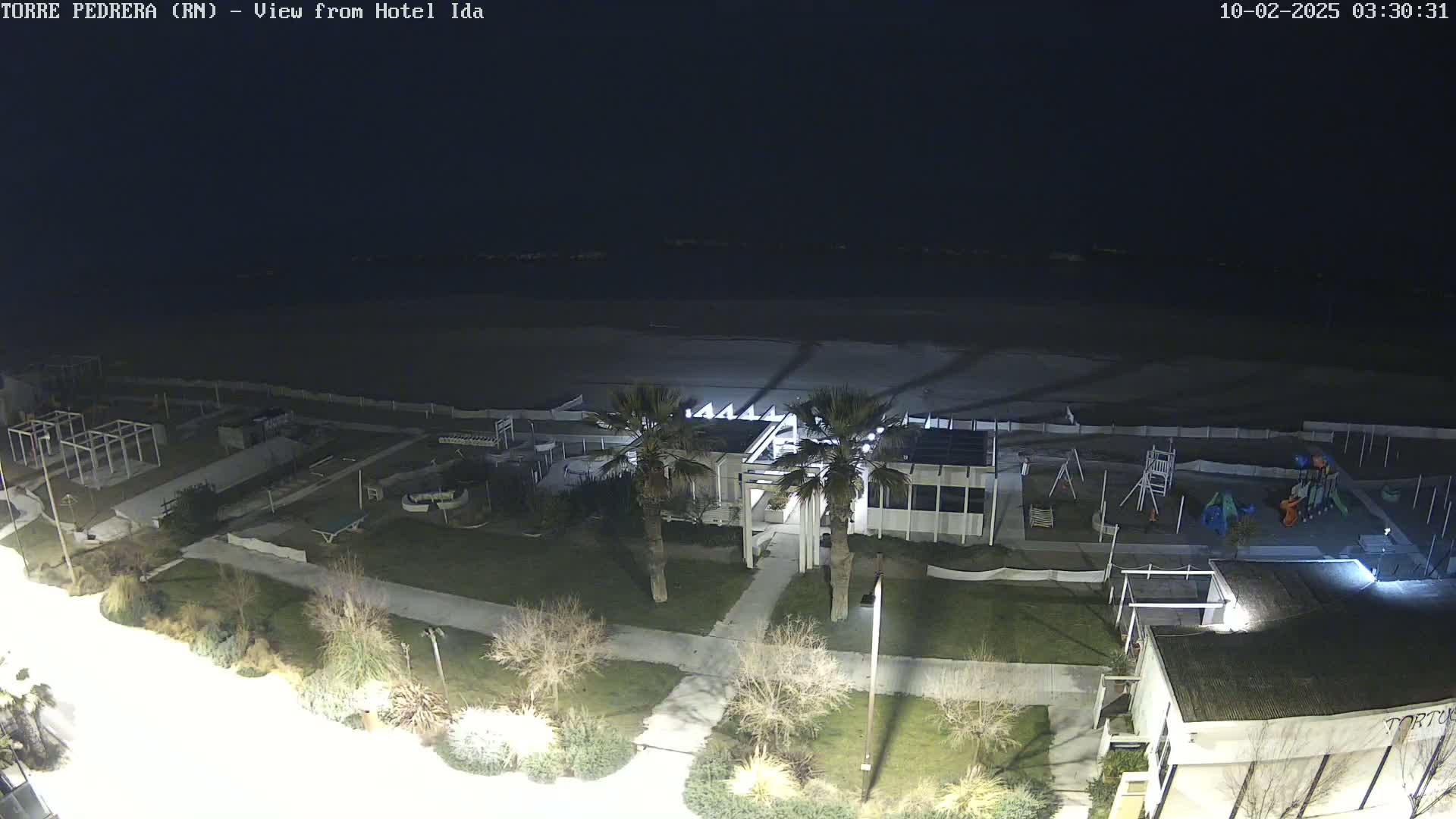 A nighttime aerial view shows a beach, a playground, and a building complex with palm trees, all illuminated by artificial light.
