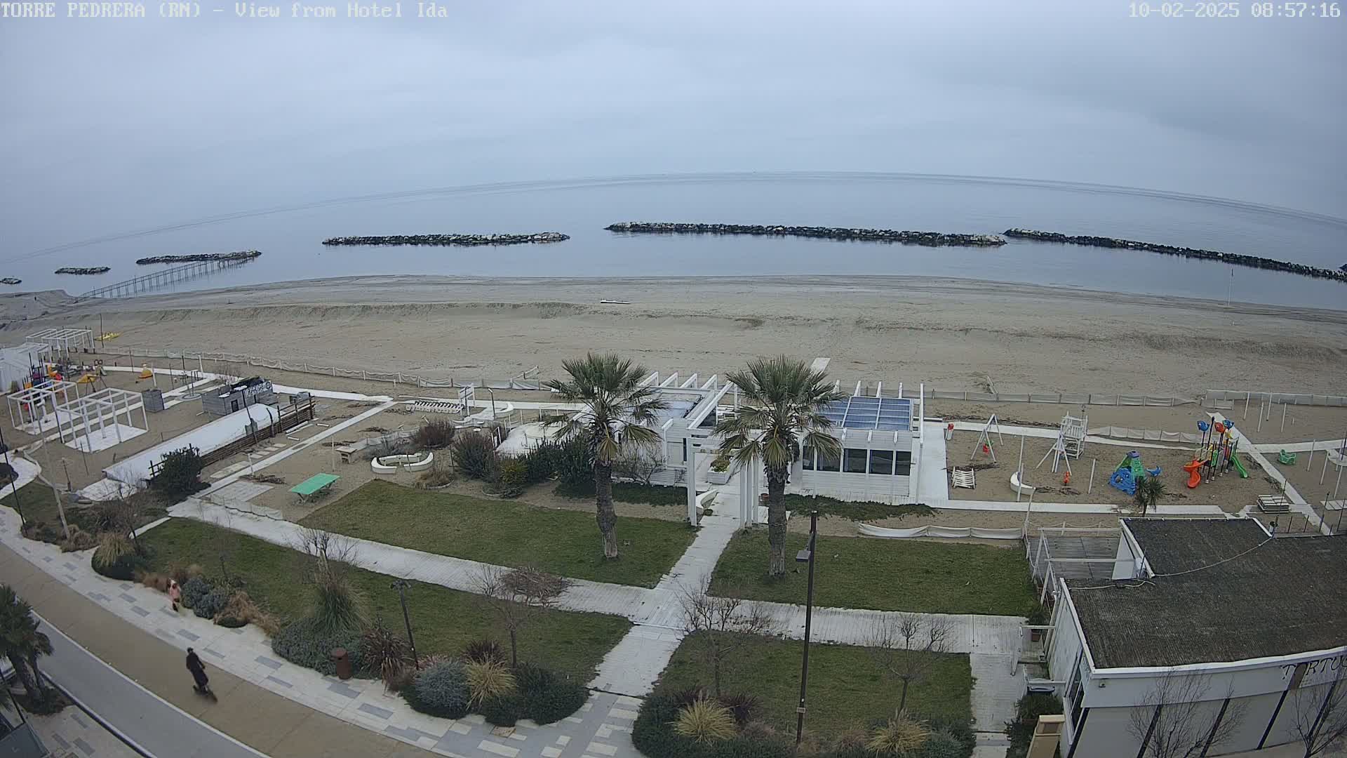 An overcast day at a beach with a sandy shoreline, a playground, a boardwalk, and several palm trees near a white building.