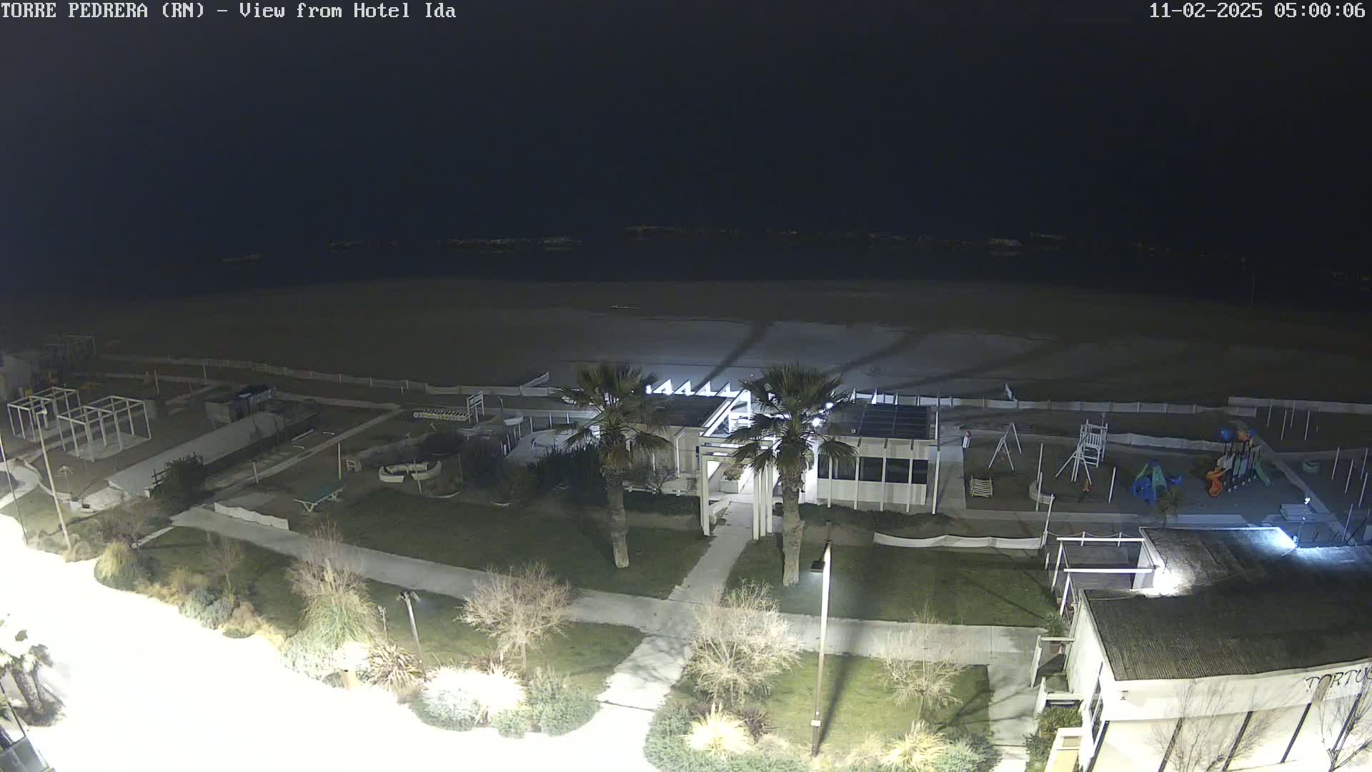 A nighttime view of a beach, showing a building, playground, and palm trees under a dark sky.