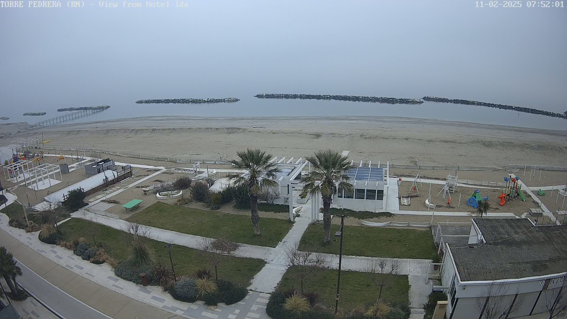 A beach scene on an overcast day shows a sandy beach, a playground, a white building with palm trees, and a calm body of water with breakwaters.