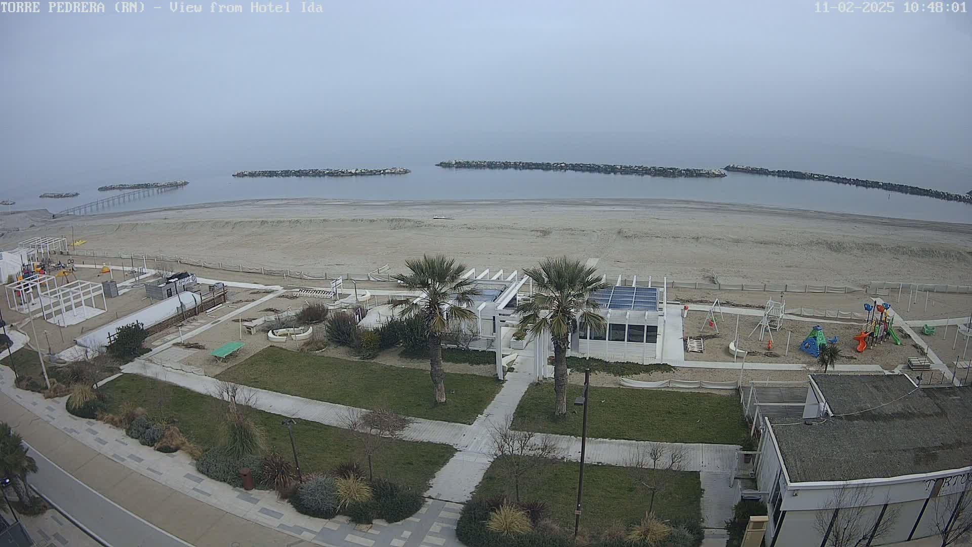 An overcast day shows a beach with a playground, a white building, palm trees, and a walkway, all near a calm body of water with breakwaters.