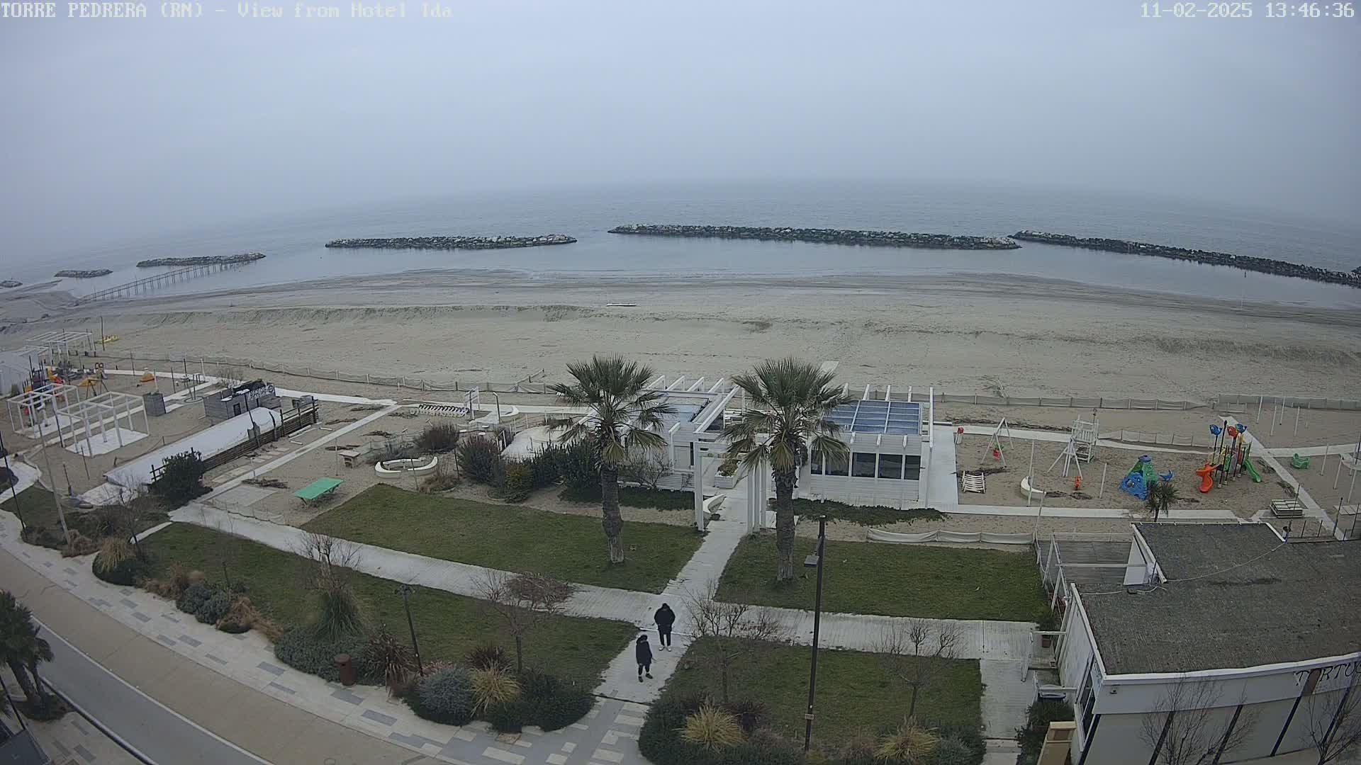 A mostly cloudy day at a beach with a sandy shore, calm water, and a beachfront area containing a playground, a restaurant, and palm trees.