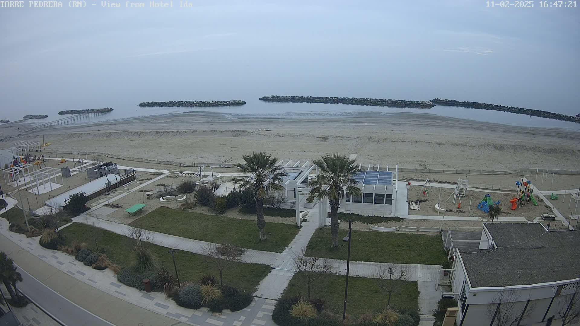 An overcast day shows a sandy beach with a playground, a white building, and palm trees near the ocean, which is calm with breakwaters visible.