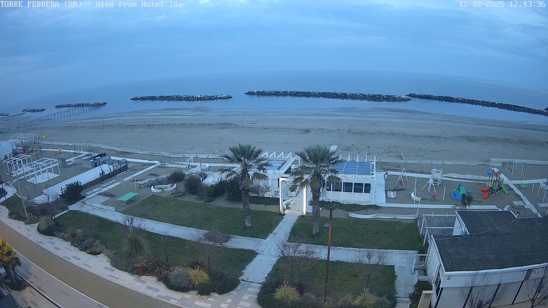 An overcast sky overlooks a sandy beach with a playground, beachfront building, and palm trees, next to a calm sea with breakwaters.
