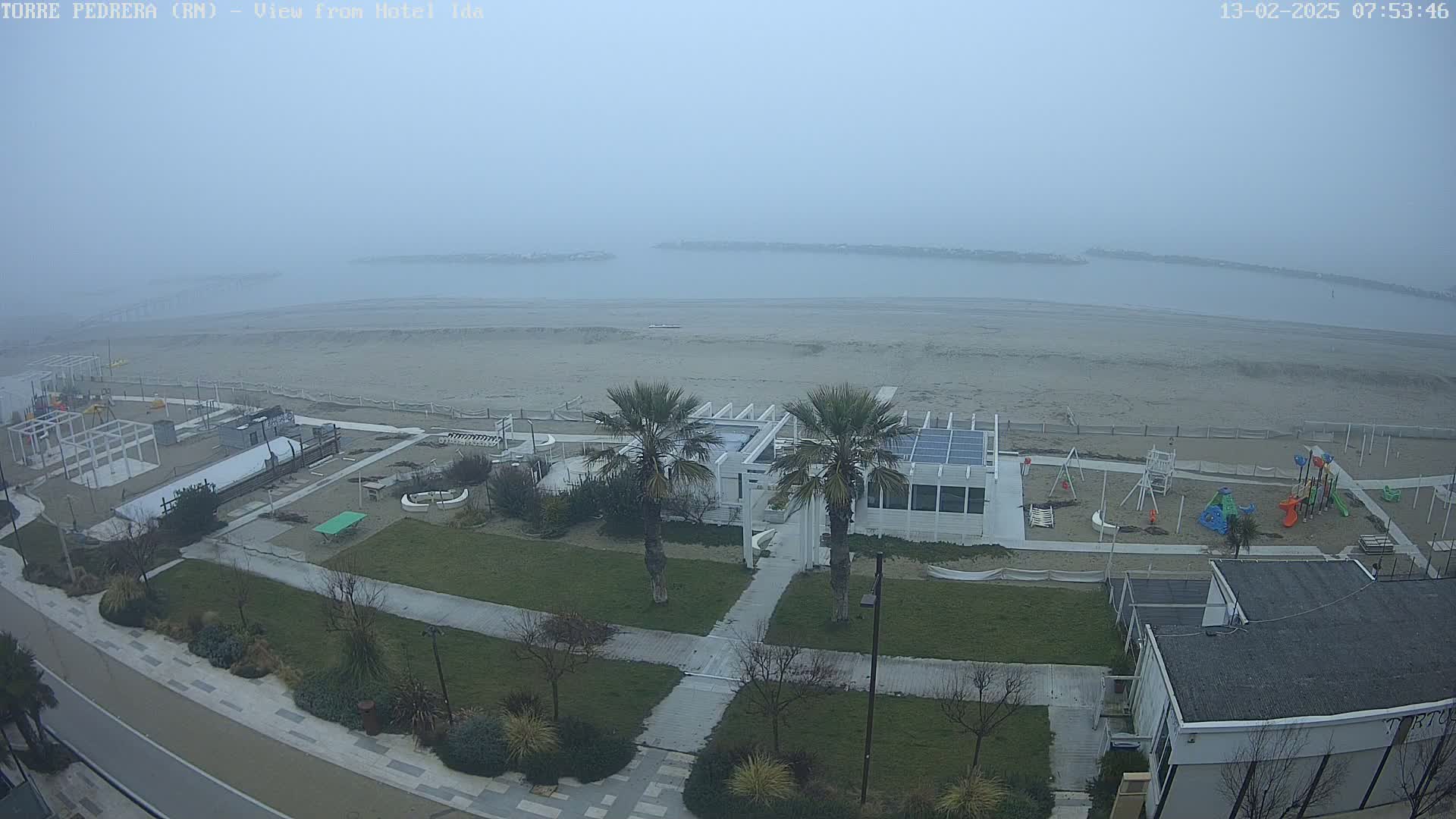 A foggy beach scene with a sandy shore, a playground, a white building with solar panels, and palm trees, viewed from above.