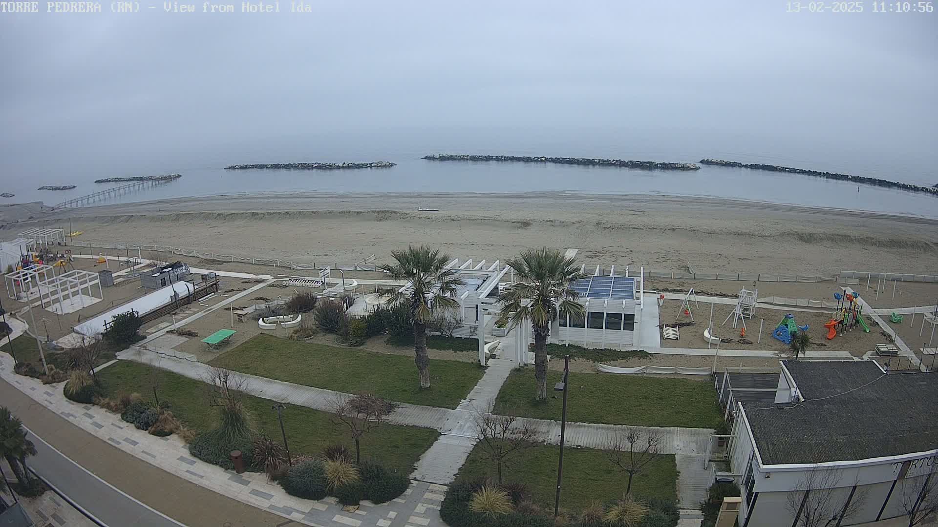An overcast day reveals a beach scene with a playground, beachfront restaurant, and palm trees, all situated near a calm sea protected by breakwaters.