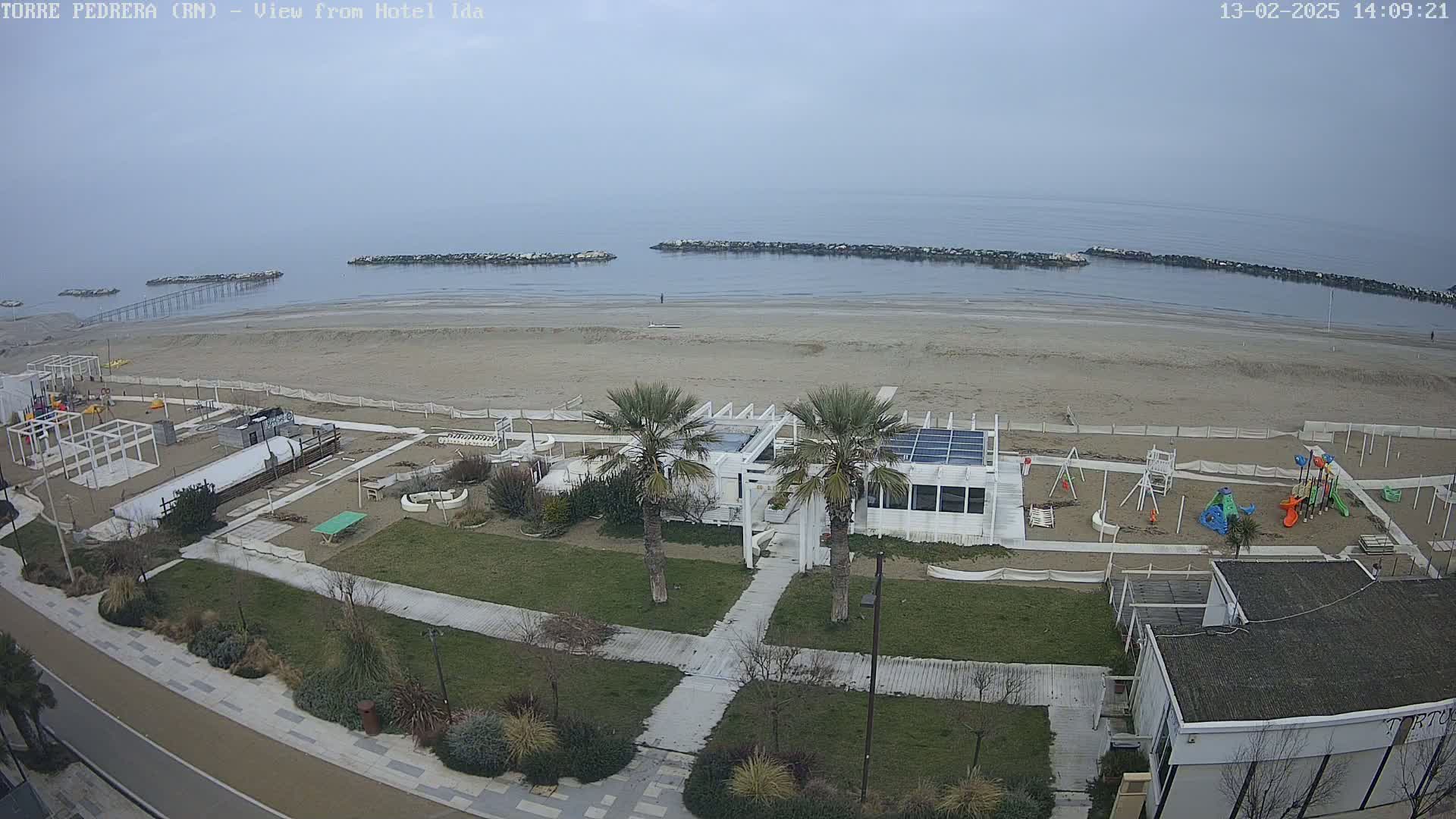 A beach with a sandy shoreline, calm water, a playground, and a white building with palm trees sits under a cloudy sky.