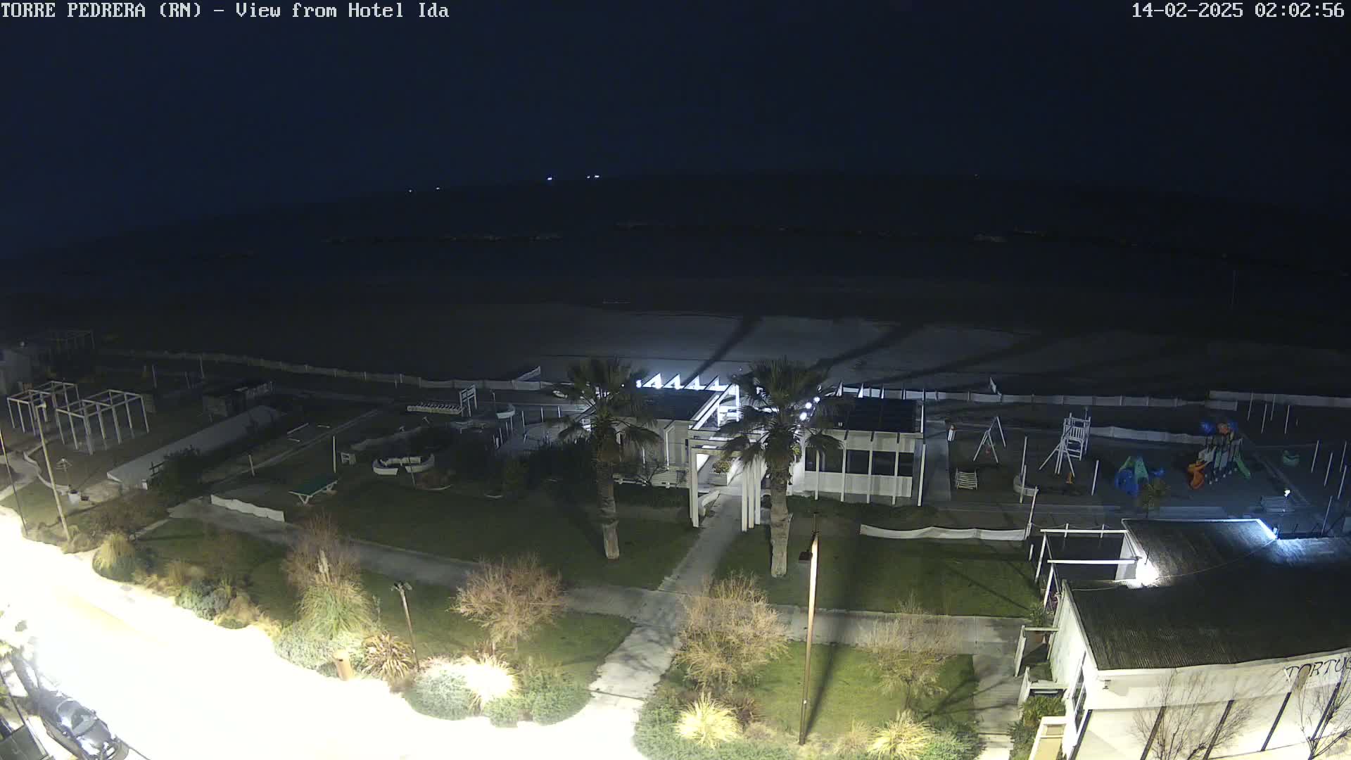 A nighttime, aerial view of a beachside area shows a building with palm trees, a playground, and an empty beach under a dark sky.