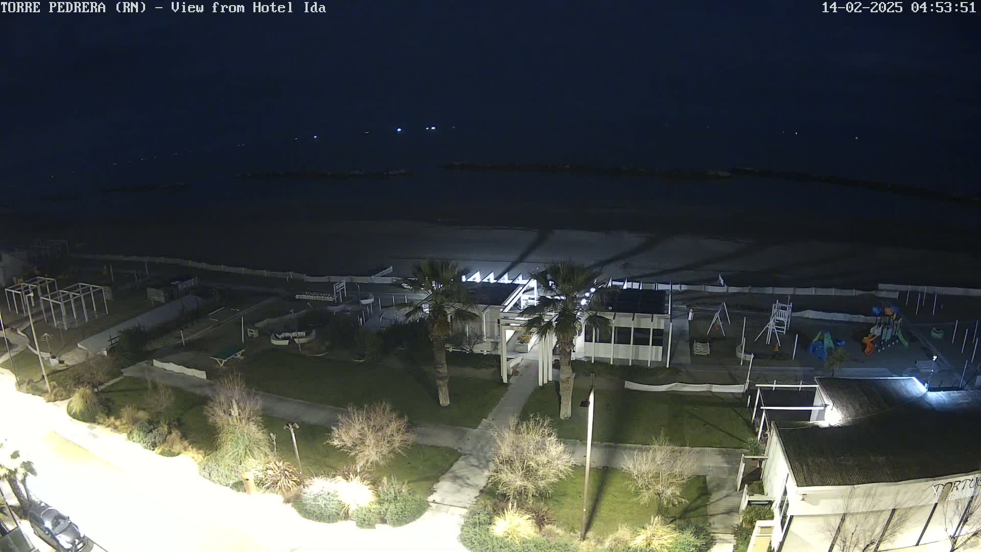 A nighttime aerial view of a beachside area shows a playground, a building with palm trees, and a dark beach under a clear, dark sky.