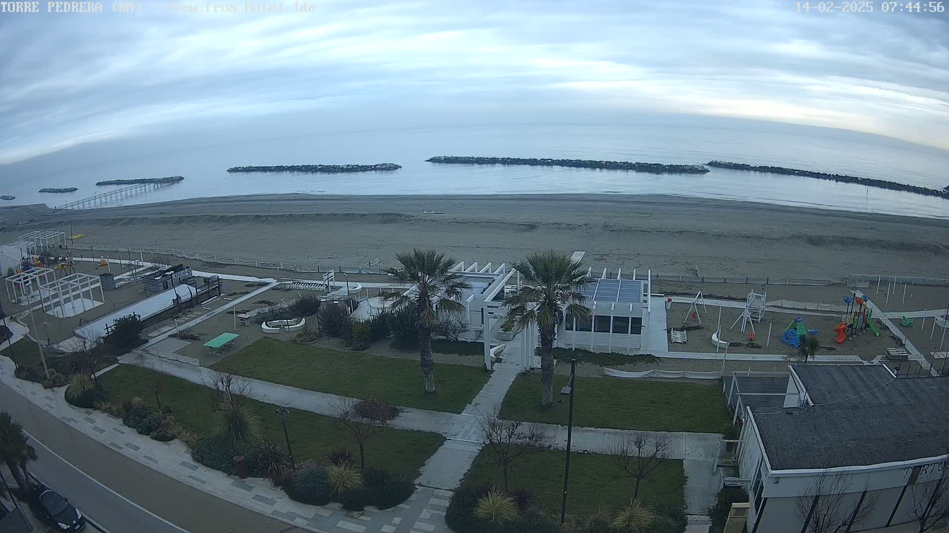 An overcast day at a beach shows a sandy shore, calm ocean water, breakwaters, a playground, palm trees, and buildings.