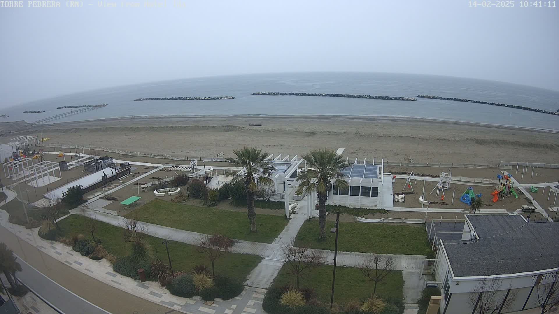 An overcast day shows a beach with a playground and beachfront building, all adjacent to a paved walkway and buildings.