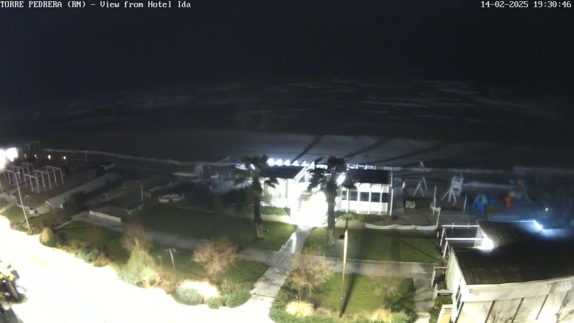 A nighttime aerial view shows a beach with dark, choppy waves under a clear, dark sky, and buildings and a playground are visible nearby.