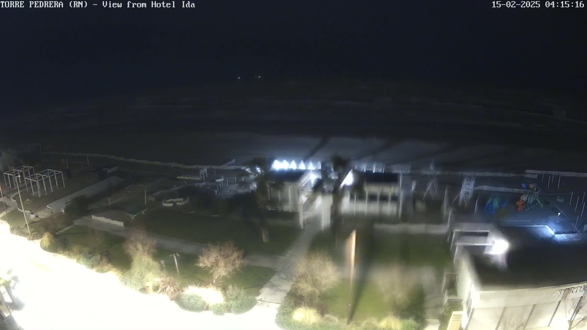 A nighttime, high-angle view shows a beach, a playground, and buildings along a shoreline under dark, cloudy conditions.