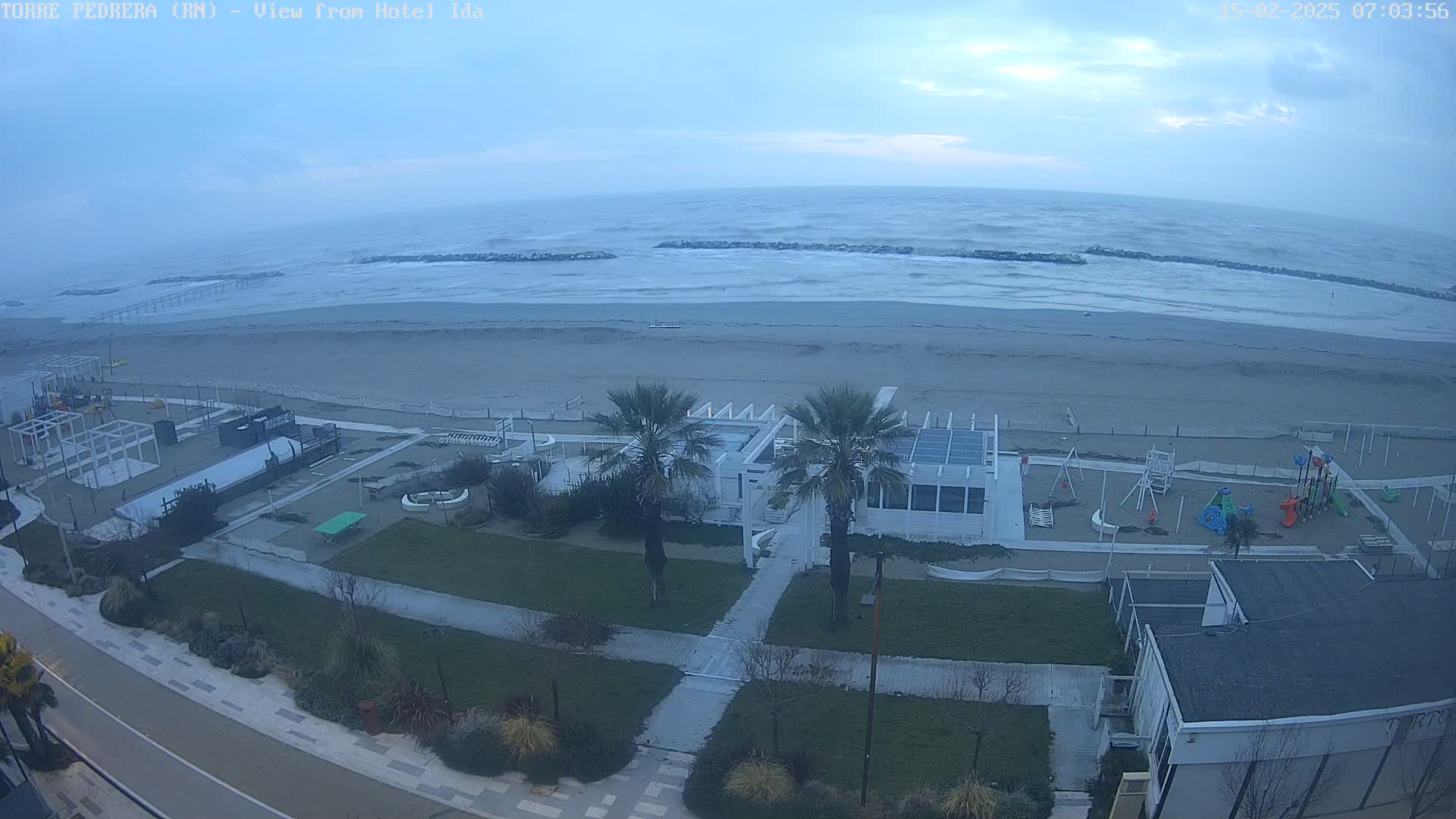 A windy, overcast day shows a beach with choppy waves, a beachfront playground, and a white building with palm trees, all viewed from above.