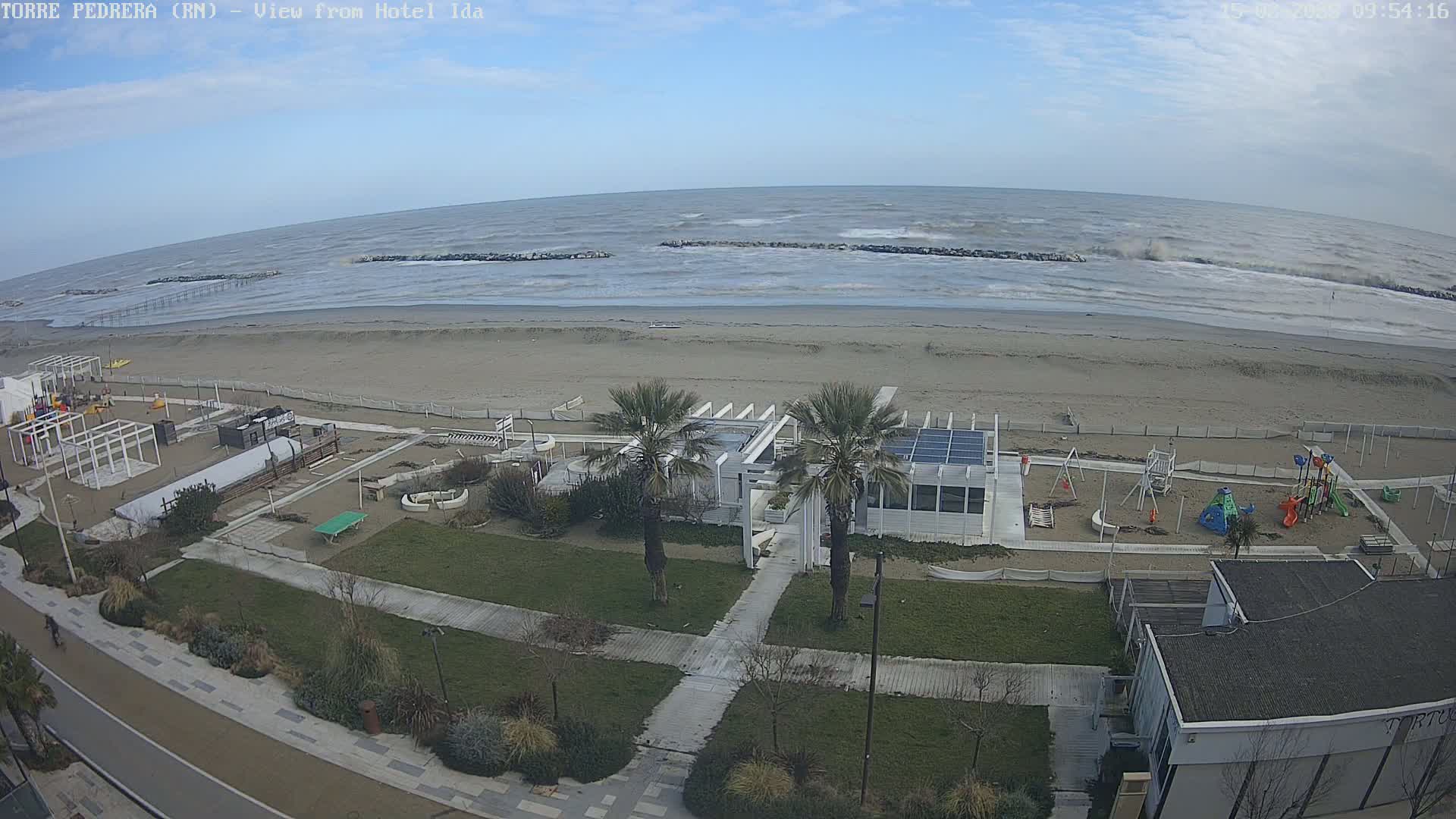 A sandy beach with a playground and a white building is visible, with a choppy sea under a mostly clear sky.