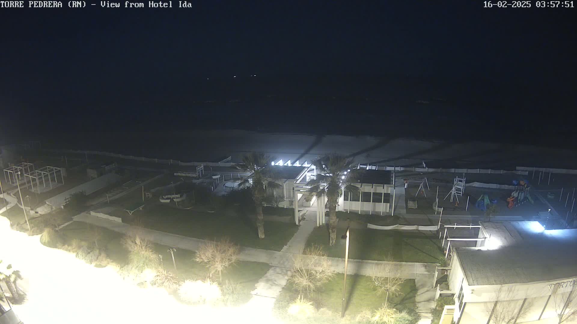 A nighttime, high-angle view of a beach, showing a mostly empty sandy shore, a playground, some buildings, and palm trees under a dark sky.
