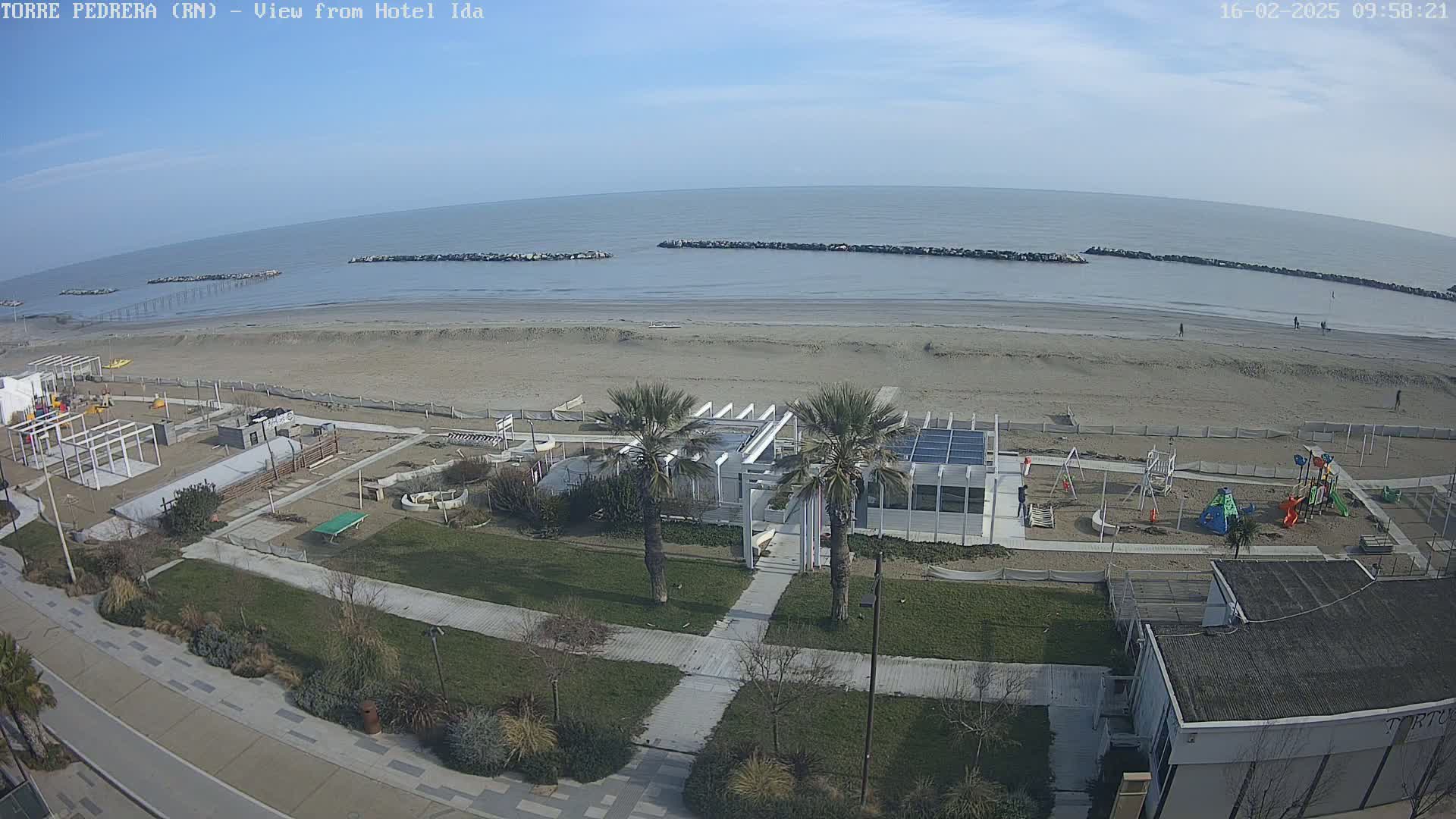 A beach scene on a mostly clear day shows a sandy beach with calm water, breakwaters, a playground, and palm trees near a building.