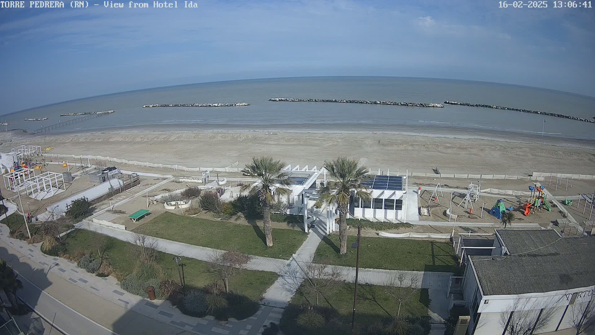 A beach with calm water under a clear blue sky, featuring a beachfront playground, a modern building with palm trees, and a paved walkway leading to the shore.