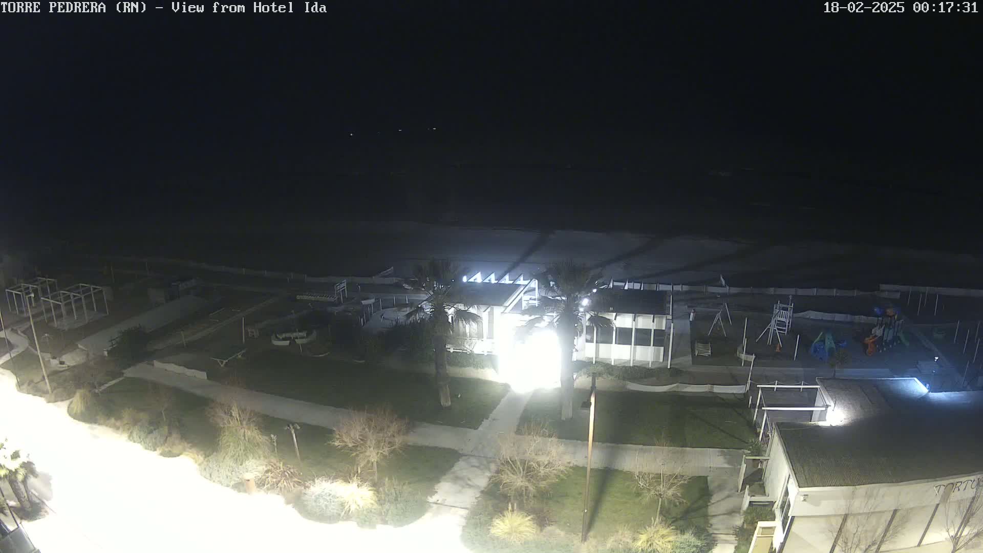 A nighttime aerial view of a beach area shows a building, playground equipment, and palm trees under a dark sky.