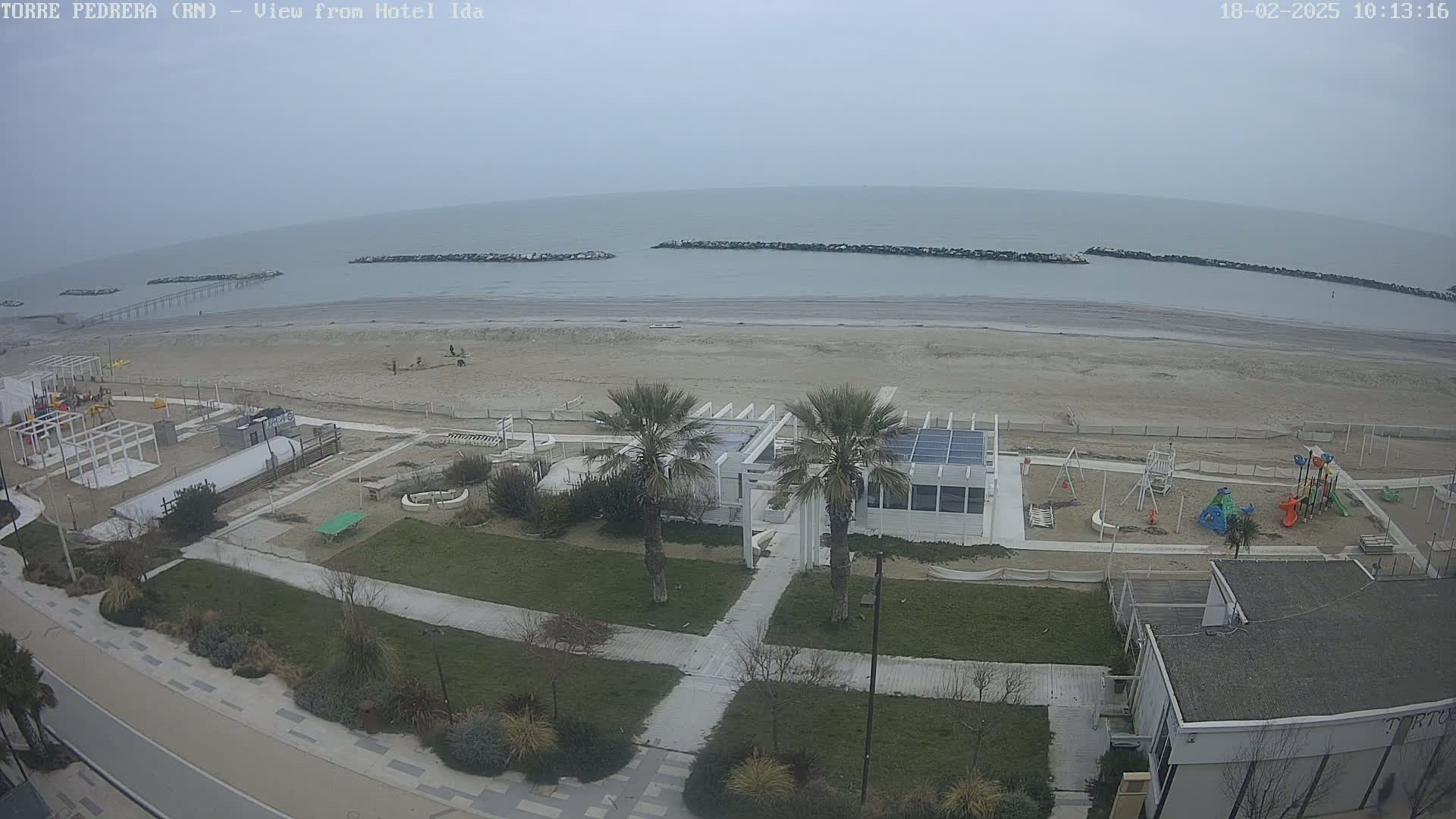 A beach with a playground and a white building is seen from above on an overcast day, with calm ocean water beyond a sandy shore protected by rock barriers.