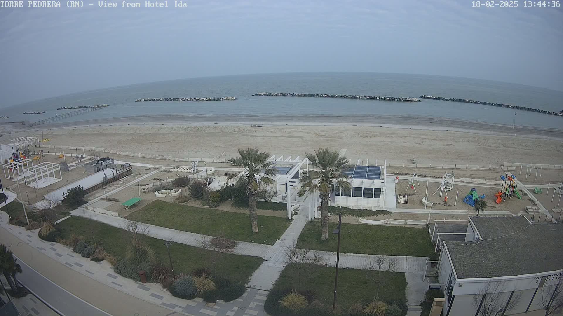 An aerial view shows a beach with calm, overcast skies, a white beachfront building with palm trees, playground equipment, and a paved walkway leading to the shore.
