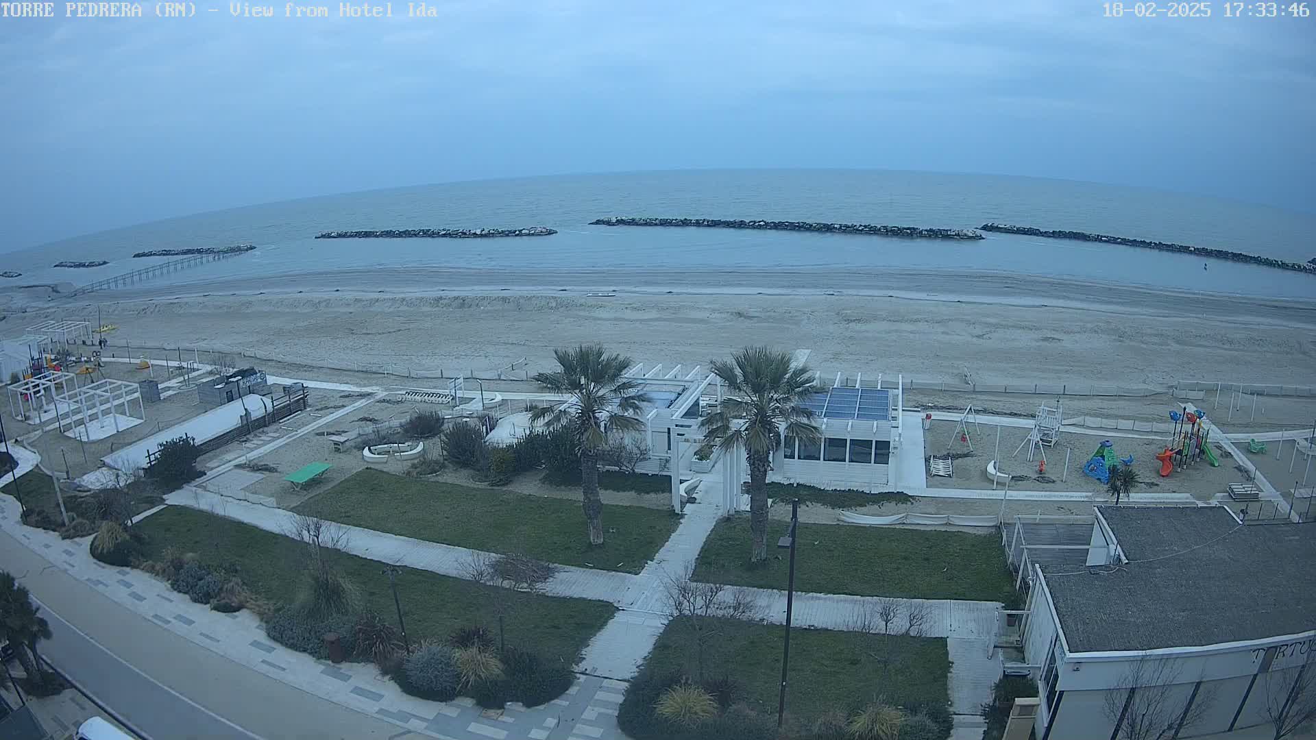 A beach with calm water under an overcast sky, featuring a beachfront building, playground, and palm trees.