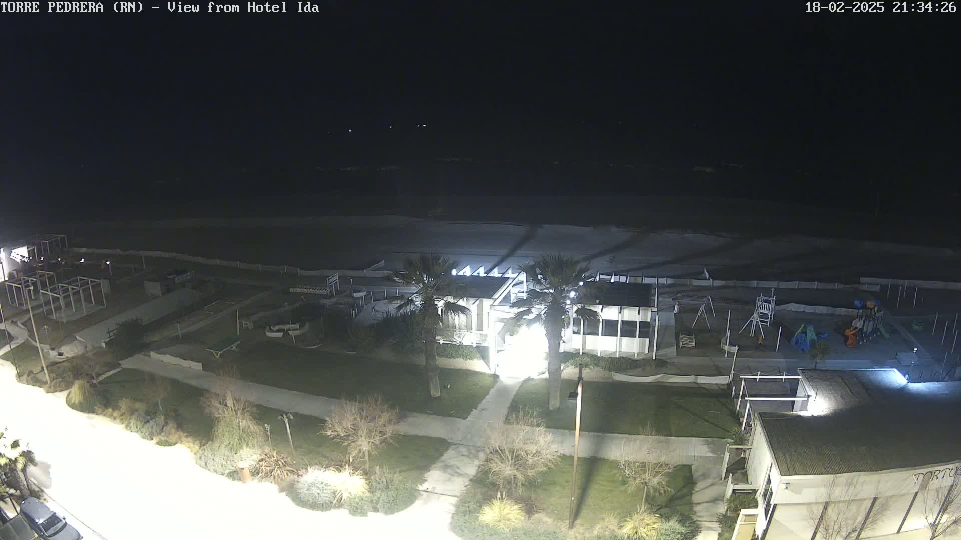 A nighttime aerial view shows a beach, a building with palm trees in front, a playground, and other structures under a dark sky.