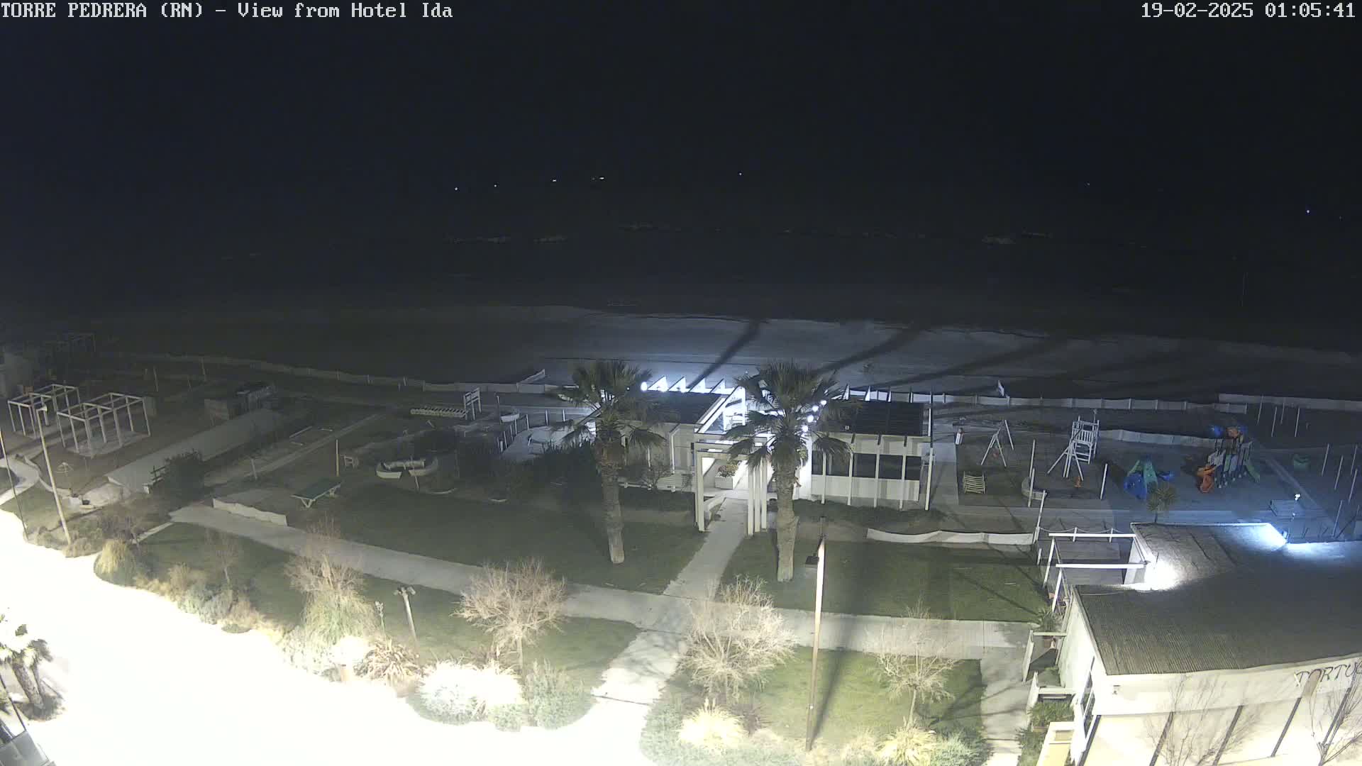 A nighttime aerial view shows a beach, a building with palm trees in front, a playground, and other structures under a dark sky.