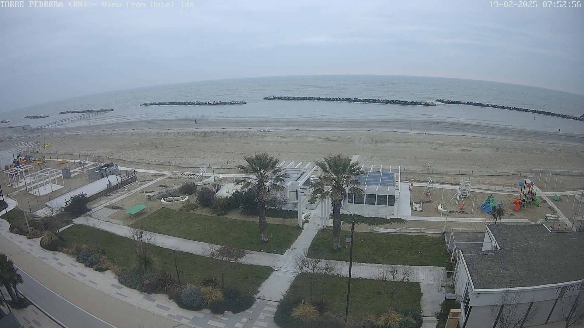 A beach with a mostly cloudy sky, featuring a sandy shoreline, calm ocean waves, a beachfront playground, and a white building with palm trees.
